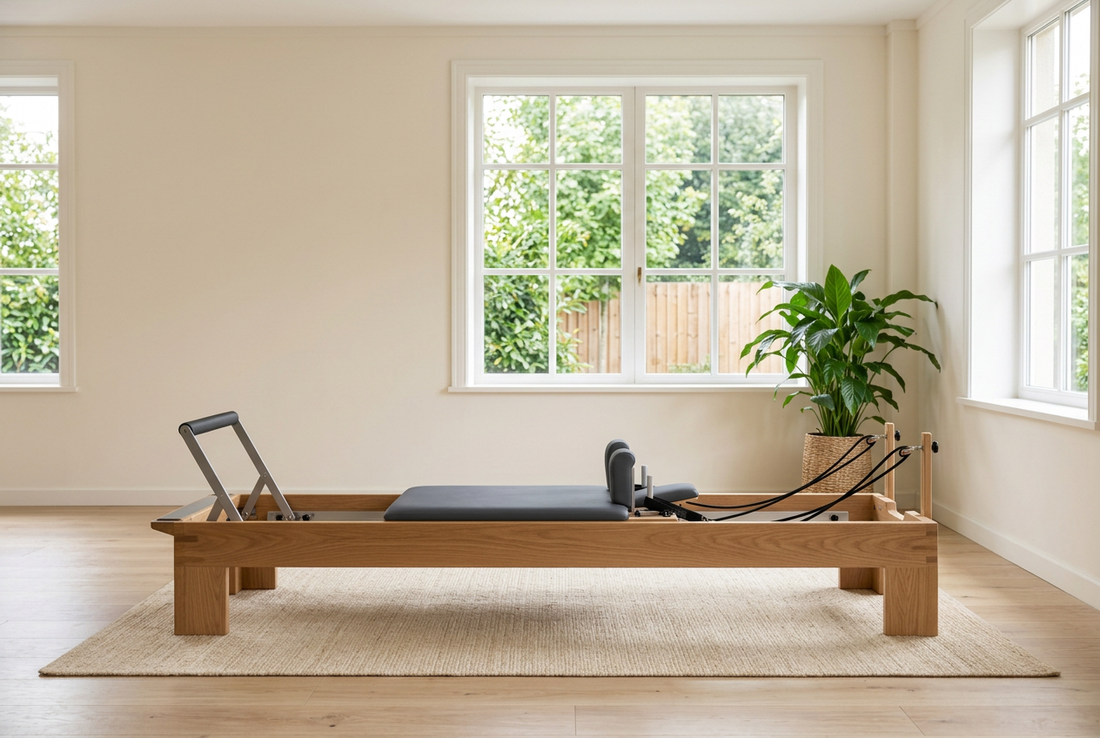 Side view of a wooden Pilates Reformer with dark padding in a bright studio with white-framed windows, leafy plants and a light wooden floor.